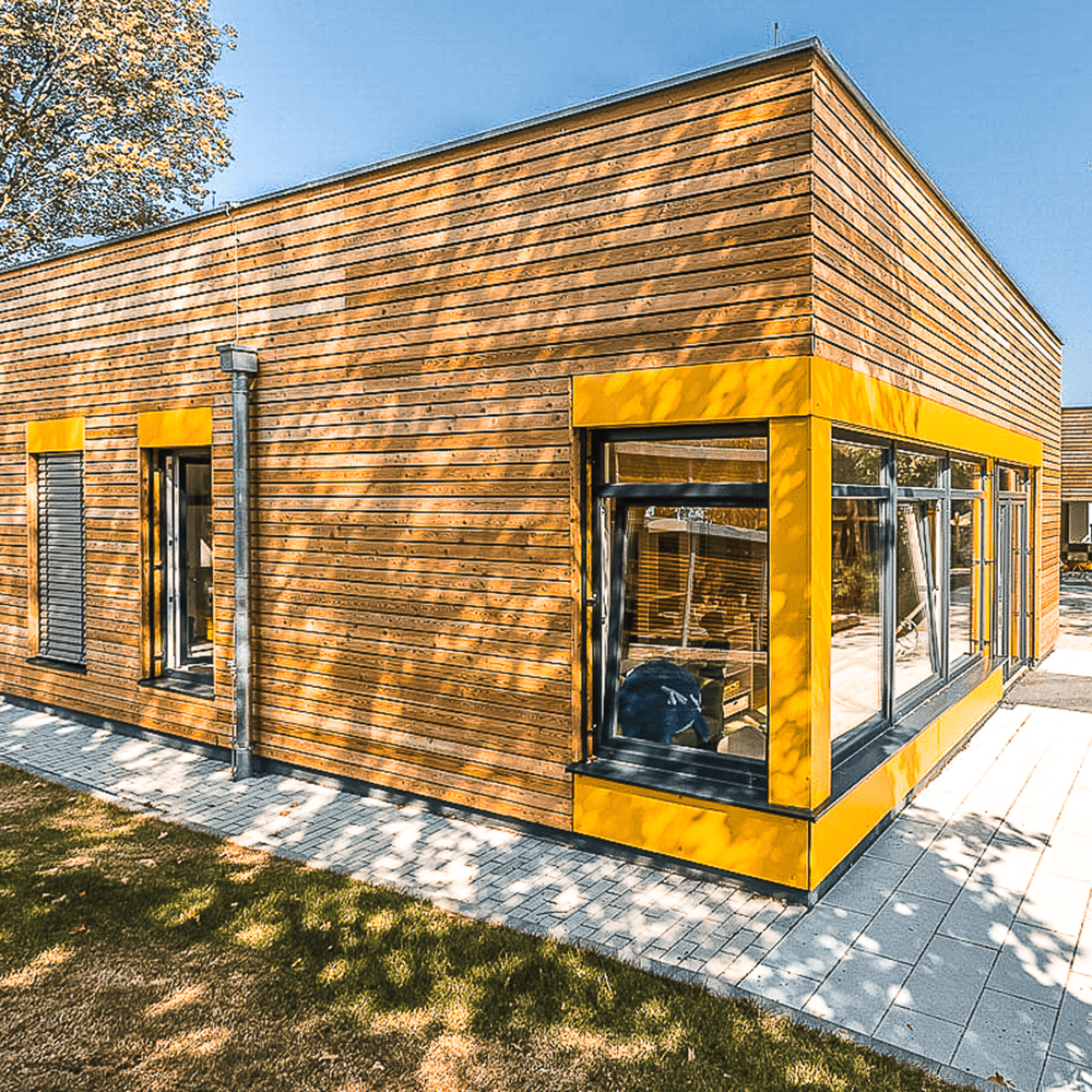 Eine Aufnahme eines Containergebäudes einer Kindertagesstätte. Die Wände sind mit Holzpaneelen verkleidet und die Fenster mit gelb lackiertem Aluminium umrahmt.