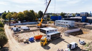 Ansicht einer Baustelle mit Tieflader und Kran, es wird eine Wohnanlage für Flüchtlinge installiert