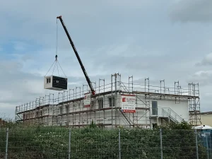 Aufnahme aus der Ferne, die ein eingerüstetes Gebäude in Modulbauweise zeigt, über das ein Modulelement per Kran gehoben wird
