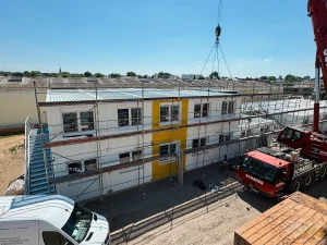 Außenaufnahme eines eingerüsteten, zweistöckigen Gebäudes zur Unterkunft für Flüchtlinge, mit einem roten Kran und einem weißen Kastenwagen davor