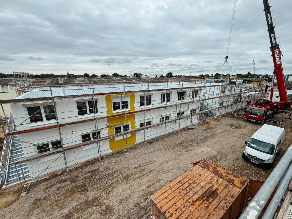 Aufnahme der Baustelle einer Wohnunterkunft für Flüchtlinge, mit einem roten Kranfahrzeug und einem weißen Kastenwagen
