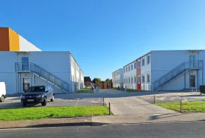 Außenansicht einer Flüchtlingsunterkunft bestehend aus mehreren weißen Modulen mit farbigen Akzenten, im Vordergrund Wiesen und Zäune, sowie ein Parkplatz mit Auto
