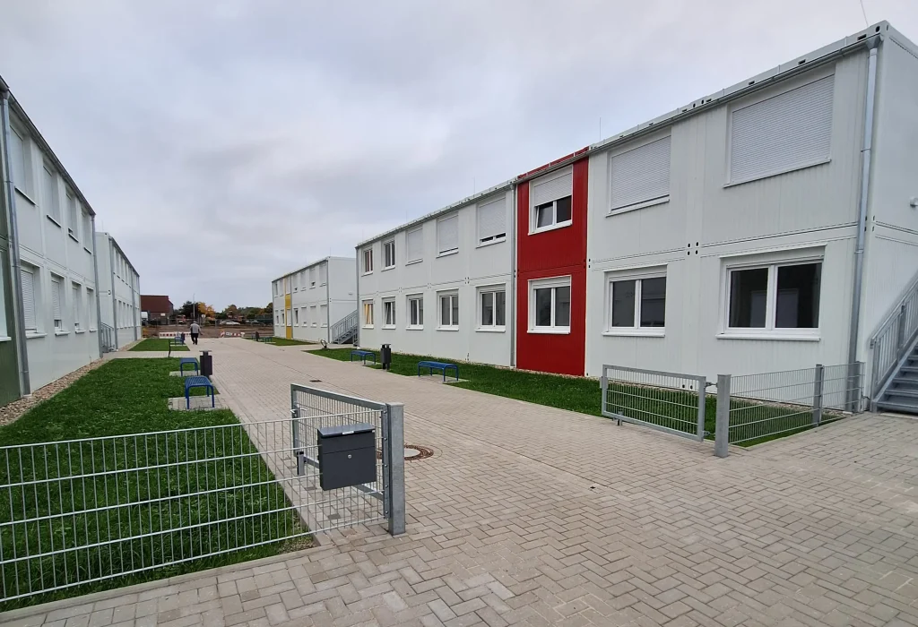 Außenansicht einer Flüchtlingsunterkunft bestehend aus mehreren weißen Modulen mit farbigen Akzenten, im Vordergrund Wiesen und Zäune