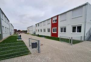 Außenansicht einer Flüchtlingsunterkunft bestehend aus mehreren weißen Modulen mit farbigen Akzenten, im Vordergrund Wiesen und Zäune