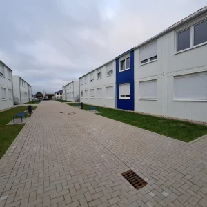 Außenansicht einer Flüchtlingsunterkunft bestehend aus mehreren weißen Modulen mit farbigen Akzenten, im Vordergrund Wiesen