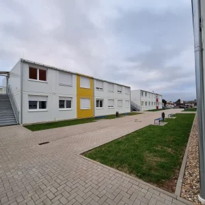 Außenansicht einer Flüchtlingsunterkunft bestehend aus mehreren weißen Modulen mit farbigen Akzenten, im Vordergrund Wiesen